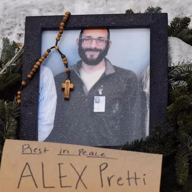 A framed photo of Alex Pretti, a man with a beard and glasses, adorned with wooden rosary beads. Below the frame, a handwritten note on cardboard reads,