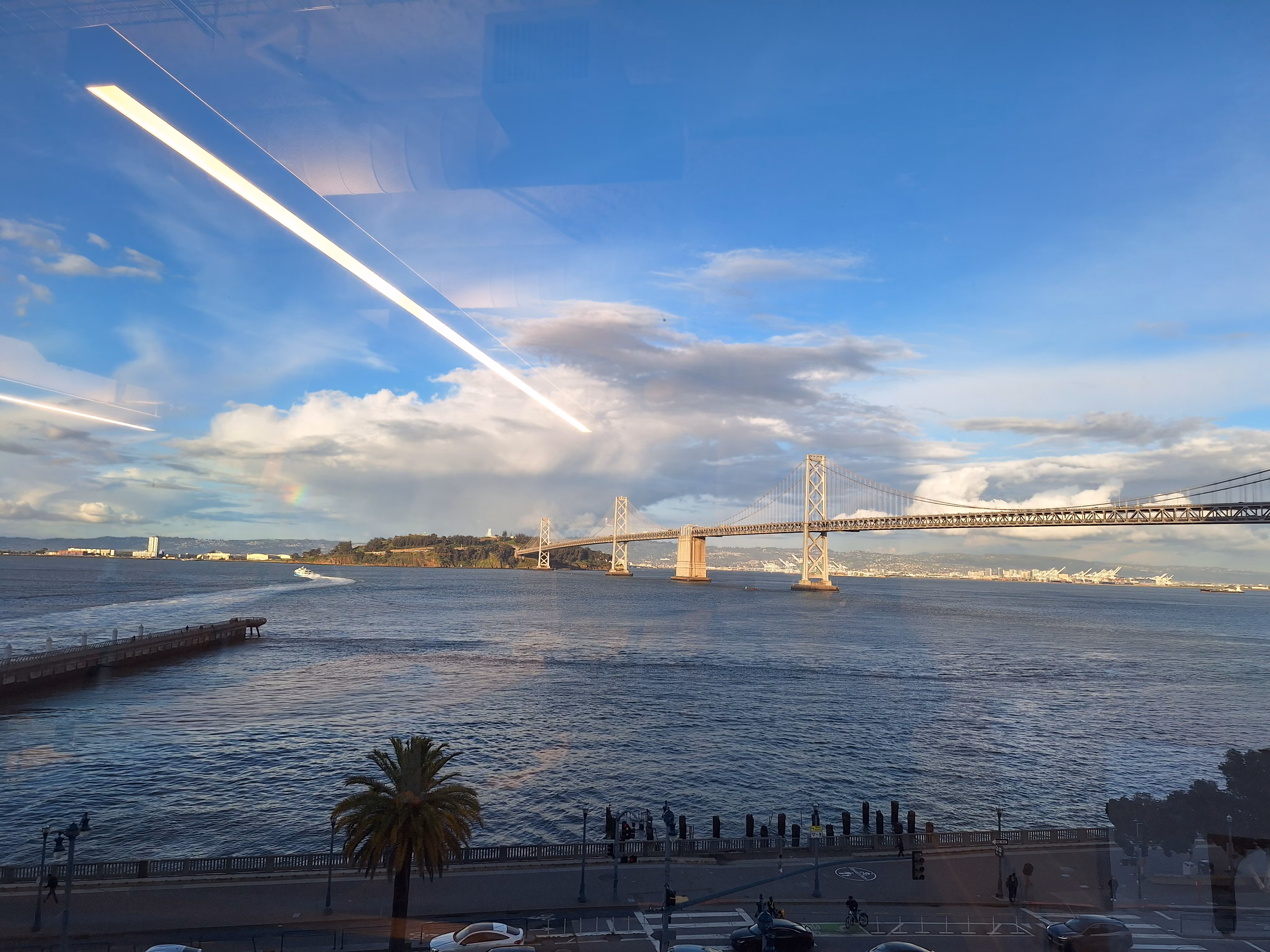The view from an office window overlooking the San Francisco–Oakland Bay Bridge, showing the blue water of the bay, a palm tree in the foreground, and a coastal road with traffic below. A small section of rainbow peeks above Treasure Island.