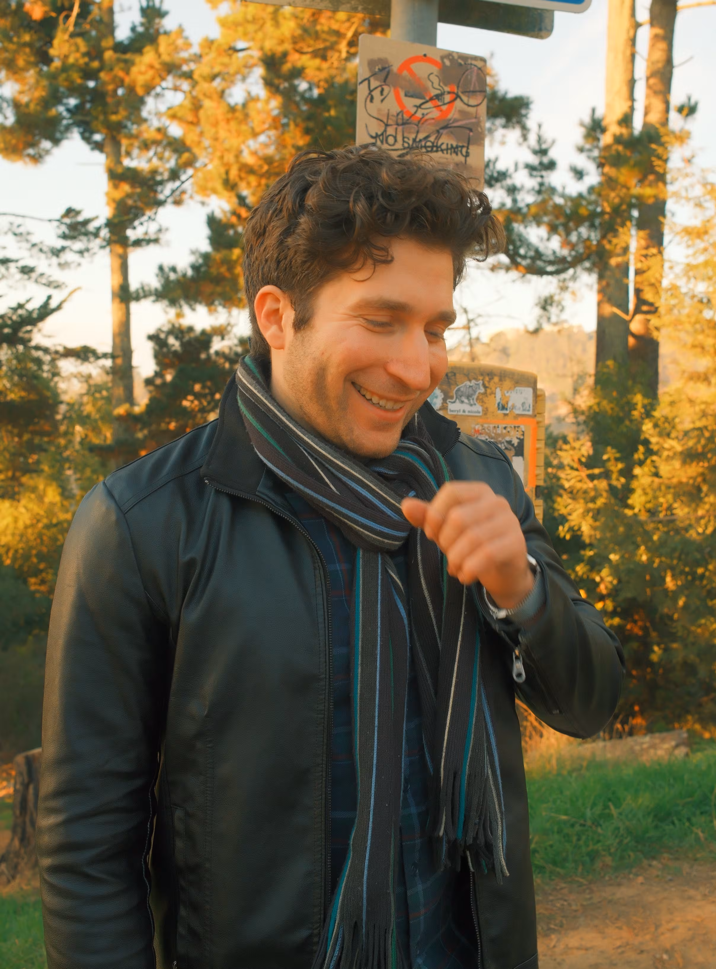 Clad in a black jacket and cold-colored scarf, Alex laughs in front of an out-of-focus yellow fall backdrop