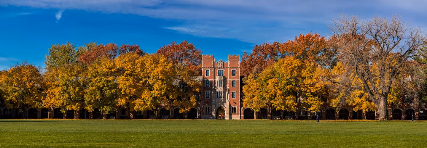 A panoramic view of the Grinnell College campus on a sunny autumn day. A large green lawn sits in the foreground before a line of trees with yellow and orange fall foliage, behind which stands a large, ornate brick building under a blue sky.