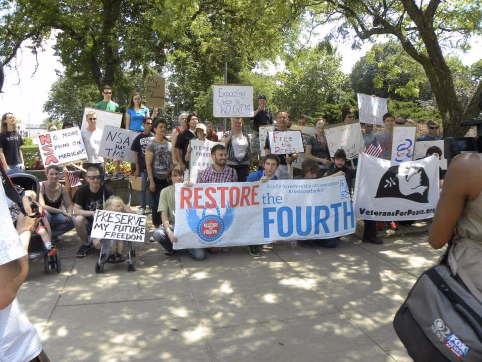 Dozens of people hold anti-spying signs in front of photographers. The author is center, helping hold a banner which reads "Restore the Fourth."