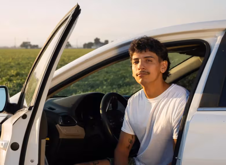 A Latino man sits in a driver's seat, door open. He faces the camera at sunset with a field behind him.