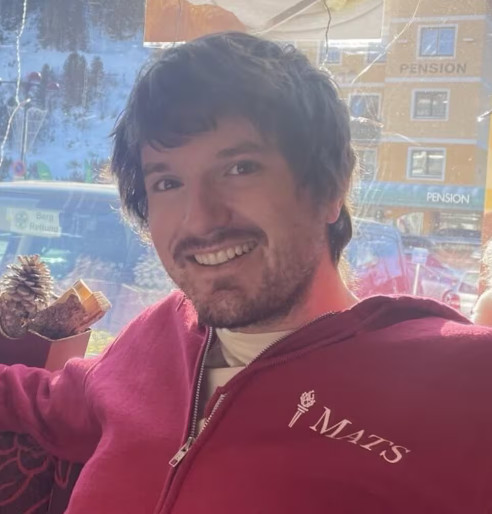 Alex Infanger smiles at the camera wearing a maroon zip-up hoodie featuring the MATS program logo.